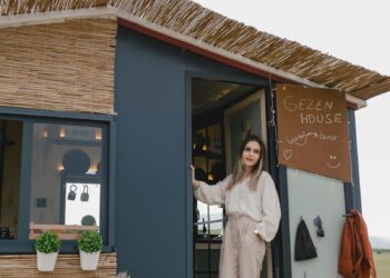Woman in boho attire standing by a mobile tiny house surrounded by nature.