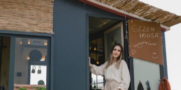 Woman in boho attire standing by a mobile tiny house surrounded by nature.
