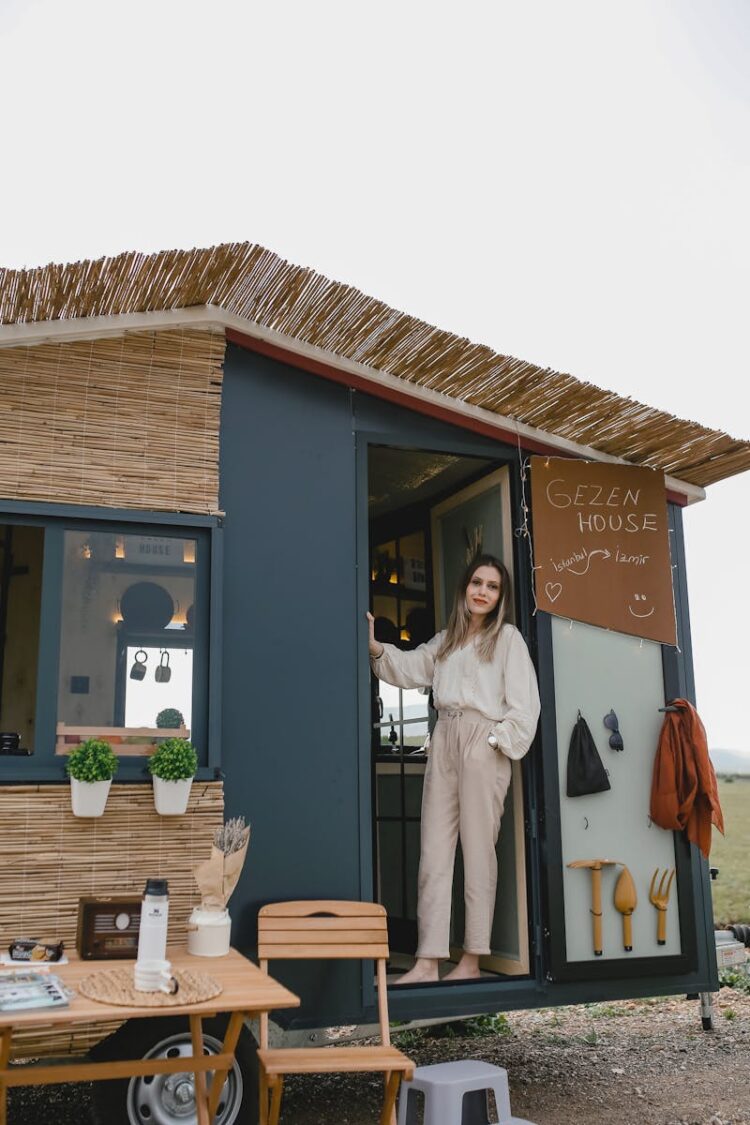 Woman in boho attire standing by a mobile tiny house surrounded by nature.