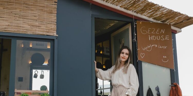Woman in boho attire standing by a mobile tiny house surrounded by nature.