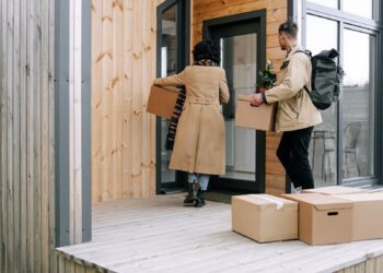 A couple carrying boxes and a plant into their new modern wooden home.