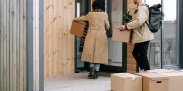 A couple carrying boxes and a plant into their new modern wooden home.