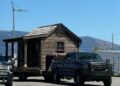 A rustic wooden cabin being towed by a pickup truck on a clear, sunny day.
