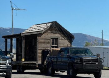 A rustic wooden cabin being towed by a pickup truck on a clear, sunny day.