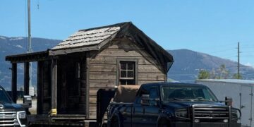 A rustic wooden cabin being towed by a pickup truck on a clear, sunny day.
