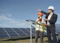 Young businessman in formal outfit and foreman in yellow vest looking away while discussing working process against solar panels