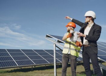 Young businessman in formal outfit and foreman in yellow vest looking away while discussing working process against solar panels