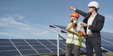 Young businessman in formal outfit and foreman in yellow vest looking away while discussing working process against solar panels