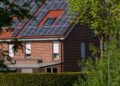 A modern brick house with solar panels on the roof, surrounded by lush greenery, under a clear blue sky.