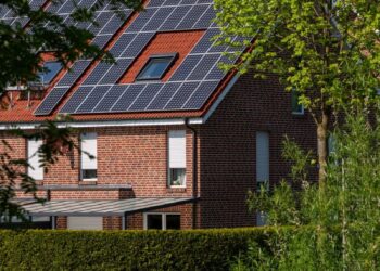 A modern brick house with solar panels on the roof, surrounded by lush greenery, under a clear blue sky.