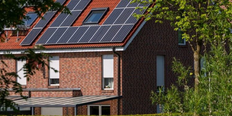A modern brick house with solar panels on the roof, surrounded by lush greenery, under a clear blue sky.