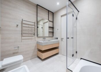 Interior of modern light bathroom with toilet and bidet next to bath and sink with faucet on cabinet under mirror