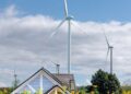 Vertical photo of wind turbines amidst sunflowers and a cottage below a blue sky.