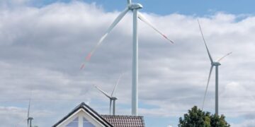 Vertical photo of wind turbines amidst sunflowers and a cottage below a blue sky.